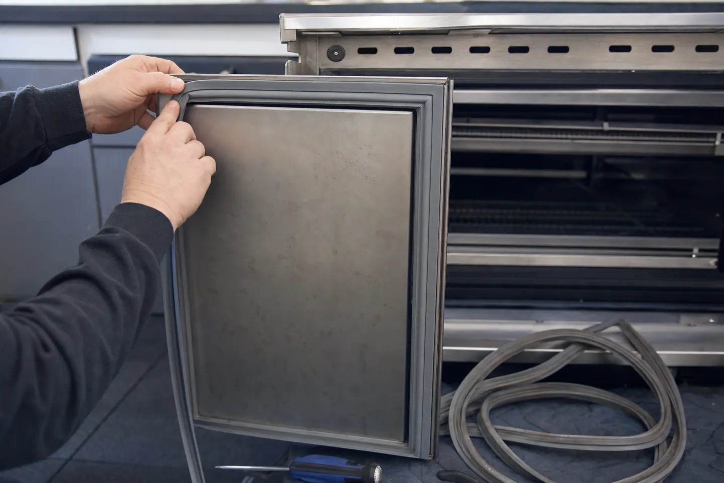 A hand holding a flexible door gasket around a small stainless door panel, the replacement gasket coiled on the floor