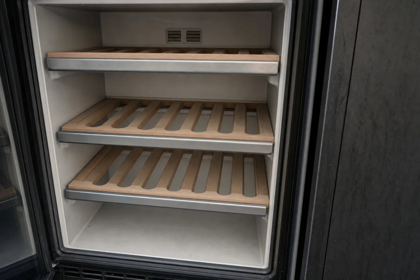 Empty undercounter cabinet interior with three slatted wooden racks on steel runners and a small vent grille set into the back wall