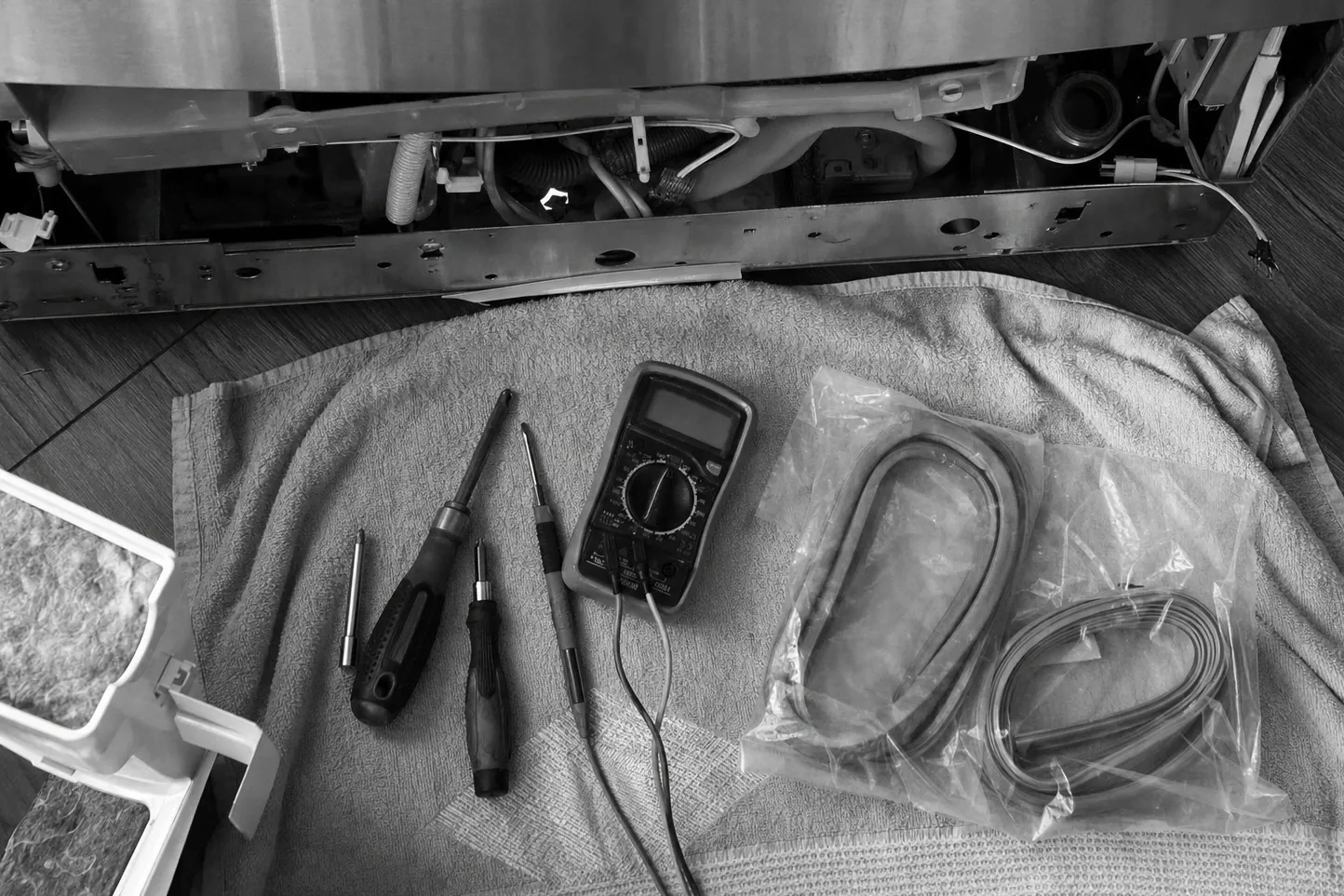 Overhead black-and-white flat-lay of three drivers, a multimeter and two bagged replacement seals on a towel, the base of a unit above with its corrugated drain hose exposed