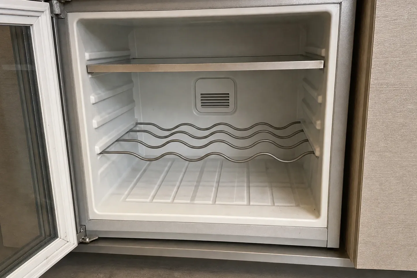 Small glass-door undercounter cabinet standing open: a plain white liner, one steel-edged shelf and a wavy wire rack above a vent grille